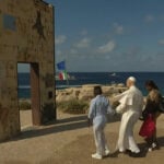 Caption: Pope Leo XIV walks hand-in-hand with African migrants through the "Gateway to Europe" monument on July 4, 2026, on the Italian island of Lampedusa. (Vatican News / Vatican Dicastery for Communication)